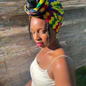 May include: A colorful head wrap with a geometric pattern in yellow, red, blue, and green. The head wrap is wrapped around the head and is paired with a cream-colored satin dress. The head wrap is a fashion accessory.