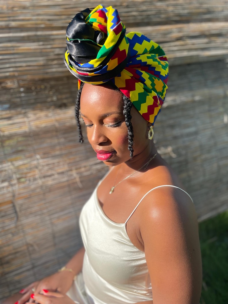May include: A colorful head wrap with a geometric pattern in yellow, red, blue, and green. The head wrap is wrapped around the head and is paired with a cream-colored satin dress. The head wrap is a fashion accessory.