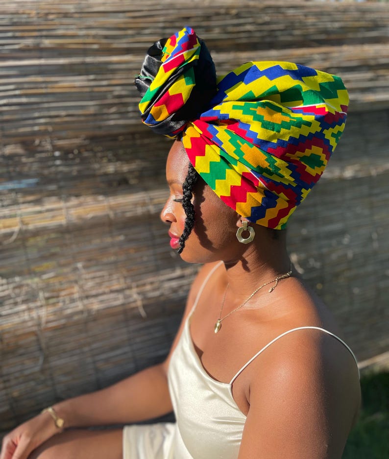 May include: A vibrant head wrap featuring a colorful geometric pattern in yellow, blue, red, and green. The head wrap is tied in a stylish knot on top, with a black satin accent. The person is wearing gold earrings and a gold necklace.
