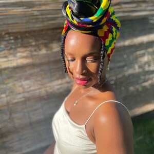 May include: A colorful head wrap with a black satin top and a vibrant pattern of yellow, green, red, and blue. The head wrap is styled on top of the head. The person is wearing a cream-colored satin dress.