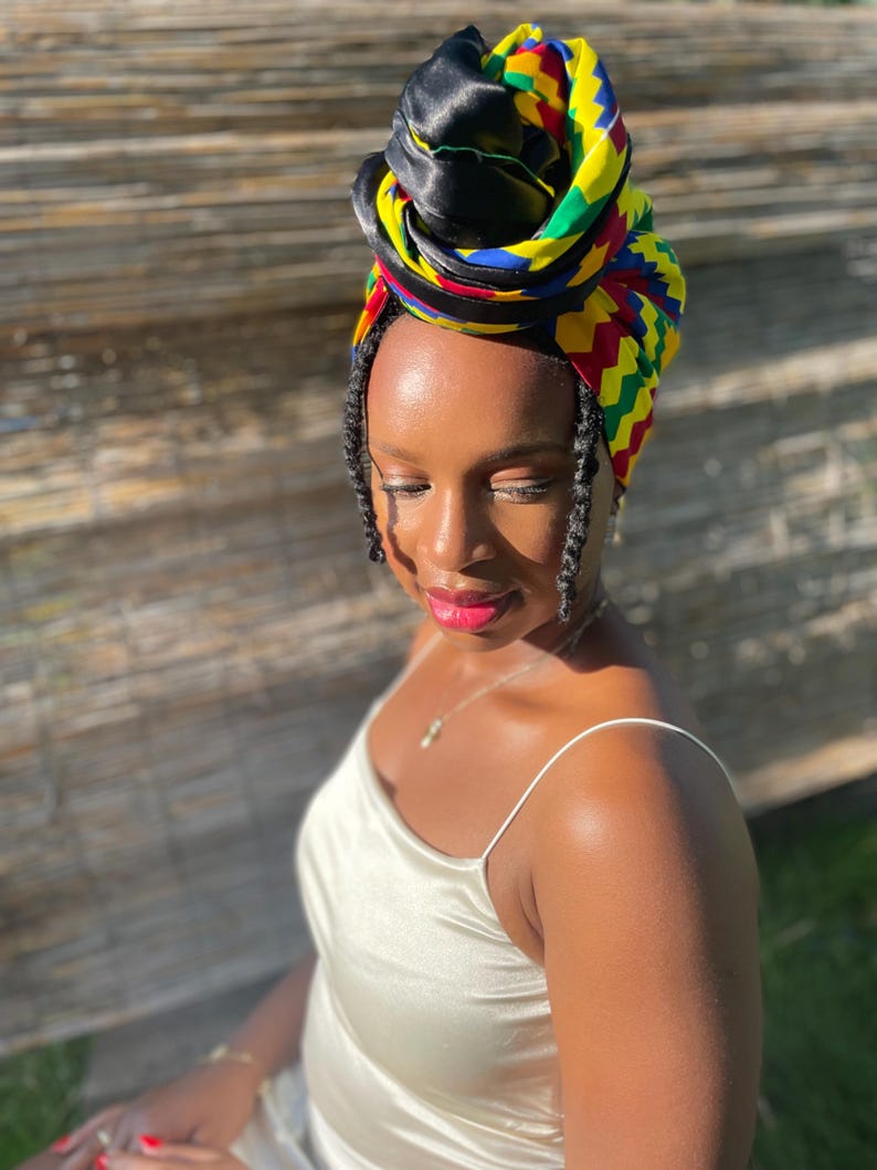 May include: A colorful head wrap with a black satin top and a vibrant pattern of yellow, green, red, and blue. The head wrap is styled on top of the head. The person is wearing a cream-colored satin dress.