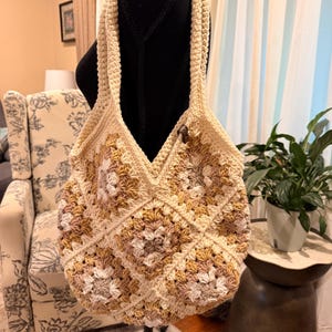 May include: A handmade crochet tote bag constructed from multiple square motifs in shades of cream, beige, and tan. The bag features a thick, textured shoulder strap and a single dark brown wooden button closure at the top center opening.