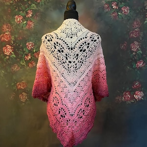 May include: A crocheted shawl with a pink and white ombre design. The shawl is displayed on a mannequin against a floral backdrop.