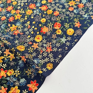 May include: A dark blue fabric featuring a dense, repeating pattern of small flowers in shades of yellow, orange, red, and light blue, interspersed with thin green stems and leaves, creating a vibrant and detailed floral surface design.