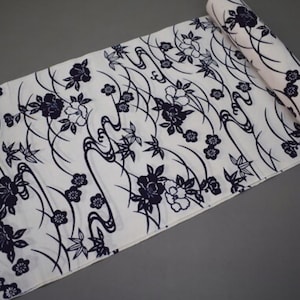 May include: White fabric with a navy blue floral pattern. The design features stylized flowers, leaves, and flowing lines. The fabric is partially rolled, revealing the pattern on the edges. The background is a solid gray.