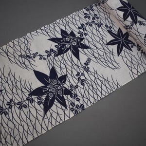 Op de afbeelding: Witte stof met een marineblauw bloemmotief met grote gestileerde bladeren en delicate wijnranken. Het ontwerp is geplaatst tegen een achtergrond van fijne, elkaar kruisende lijnen, waardoor een textuureffect ontstaat. De stof is bovenaan opgerold.