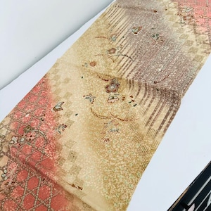 May include: A rectangular textile featuring a gradient from coral pink to beige. The fabric displays a repeating pattern of small flowers, butterflies, and geometric diamond shapes, accented with thin vertical stripes and intricate textured details.
