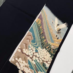 Könnte beinhalten: Schwarzer Stoff mit einem detaillierten, farbenfrohen Design. Das Design zeigt einen Kranich im Flug über einer Berglandschaft mit Kirschblüten. Die Farbpalette umfasst Türkis, Braun, Gold und Weiß.