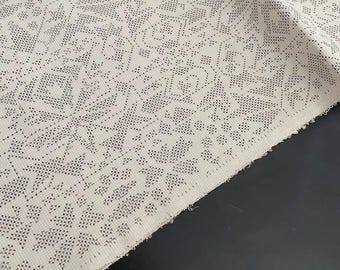 May include: Close-up of a cream-colored fabric with a detailed, repeating pattern of small, dark dots. The pattern forms geometric and floral shapes across the textile. The fabric is folded, revealing the raw edge.