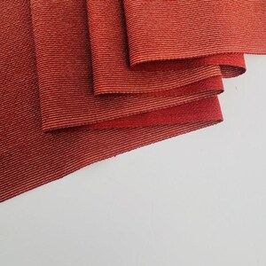 May include: A piece of fabric featuring a deep red color with a fine, horizontal ribbed texture. The material is folded in several layers, creating soft, cascading drapes that highlight the consistent linear pattern across the entire surface.