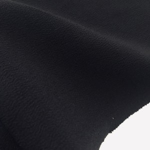 May include: Close-up of a piece of black fabric with a textured surface. The fabric is slightly wrinkled, with soft folds and shadows. The material appears to be a medium-weight textile, suitable for clothing or home decor.