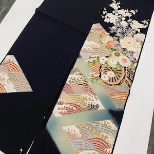 Könnte beinhalten: Schwarzer Kimono-Stoff mit farbenfrohen Wellen- und Blumenmustern. Das Design umfasst weiße, rosa und pfirsichfarbene Blumen sowie detaillierte Wellenmuster in Braun-, Blau- und Goldtönen. Der Stoff liegt auf einer weißen Oberfläche.