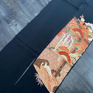 Könnte beinhalten: Ein schwarzes Textil mit einem detaillierten, farbenfrohen Muster. Das Design umfasst orange, rote und weiße florale Elemente sowie ein Wellenmuster. Das Textil ist wahrscheinlich Teil eines größeren Kleidungsstücks oder Dekorationsobjekts.