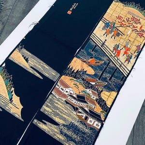 Könnte beinhalten: Zwei Stücke dunkelblauen Stoffes mit japanischem Druck. Ein Abschnitt zeigt eine Flussszene mit Booten und Figuren, der andere eine Berglandschaft. Das Kunstwerk weist goldene, braune und rote Akzente auf.
