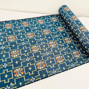 May include: A roll of fabric featuring a deep blue background with a repeating geometric pattern of yellow lines and white and orange floral-like motifs. The textile displays a woven texture with a consistent grid layout across the entire surface.