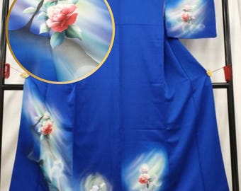May include: A vibrant blue kimono with floral artwork. The design features a large, detailed illustration of a red and white flower with green leaves. Additional smaller floral designs are scattered across the garment, creating a flowing, artistic aesthetic.