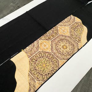 Könnte beinhalten: Schwarzer Stoff mit einer dekorativen Mittelbahn. Die Bahn hat ein sich wiederholendes geometrisches Muster in Gold, Braun und Creme, mit einem floralen Design in jeder Form. Die Kanten der Bahn sind mit einem cremefarbenen Rand versehen. Der Stoff ist wahrscheinlich für ein Kleidungsstück.
