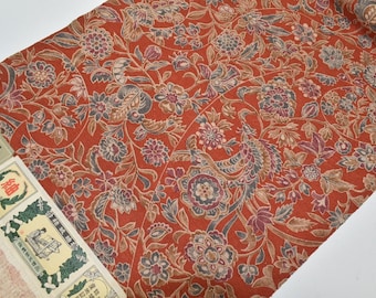 May include: A close-up of a red fabric with a floral pattern. The fabric has a paisley design with brown, blue, and pink flowers. The fabric is folded in half and the edge is visible.