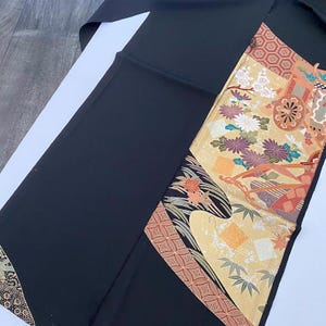Könnte beinhalten: Schwarzer Kimono mit einem dekorativen Panel mit floralen und geometrischen Mustern in Gold-, Orange-, Lila- und Grüntönen. Der Kimono ist aus einem glatten, dunklen Stoff gefertigt.