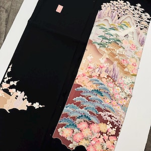 Könnte beinhalten: Ein schwarzer Stoff mit einem detaillierten, farbenfrohen Blumen- und Landschaftsdesign. Das Design zeigt rosa Kirschblüten, grüne Kiefern und eine Berglandschaft mit einem Kranich. Der Stoff hat eine rechteckige Form.