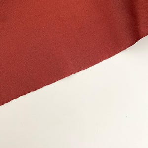 May include: A close-up of a section of red-brown fabric with a textured weave. The fabric is diagonally positioned against a white background, with a raw, unfinished edge visible.