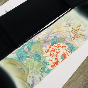 Könnte beinhalten: Ein Abschnitt eines Kimonos mit schwarzem Rand und detailliertem Blumen- und Vogeldesign. Das Design zeigt eine große weiß-orangefarbene Blume, grüne Blätter und einen Vogel mit gelben, braunen und roten Federn. Der Hintergrund ist hellgrün und weiß.