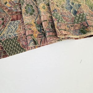 May include: A piece of woven fabric featuring a dense, multi-colored pattern of floral motifs, geometric shapes, and swirling lines in shades of dusty rose, sage green, mustard yellow, and deep blue, with a textured, tapestry-like appearance.