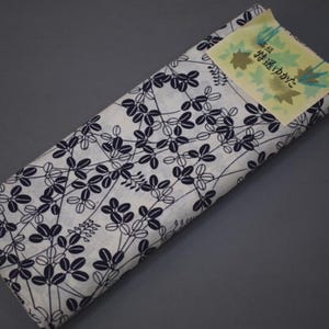 May include: Folded white fabric with a navy blue leaf pattern. A small yellow tag with Japanese text and leaf illustrations is attached to the fabric. The fabric is folded and appears to be new.