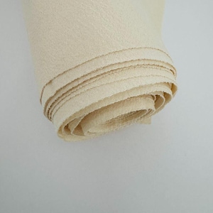 May include: A tightly rolled bolt of off-white, cream-colored fabric with a visible woven texture. The material features a plain weave construction with raw, slightly frayed edges visible along the spiral layers of the roll.