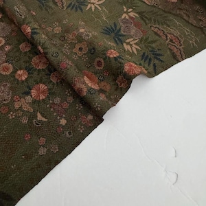 May include: A textured olive green fabric featuring a repeating pattern of small flowers, leaves, and botanical elements in shades of pink, dark blue, and muted brown. The textile has a visible weave and is draped to show its flexible, folded form.