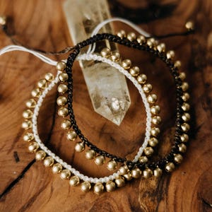 Può includere: Due braccialetti con perline dorate su cordini neri e bianchi. I braccialetti sono su una superficie di legno.