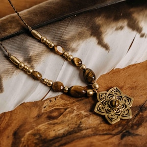 Può includere: Una collana artigianale con perline di occhio di tigre e un ciondolo a forma di fiore in ottone. La collana è infilata su un cordoncino marrone e poggia su una piuma e una superficie di legno. Il ciondolo ha un design intricato.