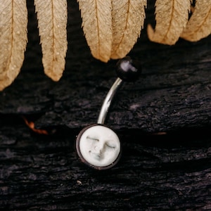 Puede incluir: Un piercing de ombligo plateado con un dije de luna blanco y negro. El dije de luna tiene una cara con los ojos cerrados.