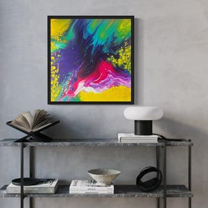 Könnte beinhalten: Abstraktes Gemälde mit leuchtenden Farben in Gelb, Grün, Blau, Lila und Rosa. Das Gemälde ist in Schwarz gerahmt und auf einem Regal mit Büchern und einer Lampe ausgestellt.