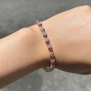 May include: A gold bracelet featuring alternating red and clear gemstones in a floral design. The bracelet is displayed on a wrist.