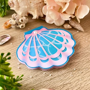 Puede incluir: Una manualidad de papel con forma de concha marina en rosa y azul, con un acento de perla. La manualidad está rodeada de conchas marinas, flores artificiales y vegetación, creando un tema playero. La manualidad tiene un diseño en capas con un interior azul claro.