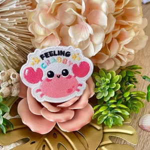 Puede incluir: Un parche bordado blanco con un cangrejo rosa, pinzas en forma de corazón y el texto "Feeling Crabby". El parche tiene un borde blanco y estrellas decorativas. Se encuentra sobre una rosa rosa con un fondo floral y una hoja dorada.