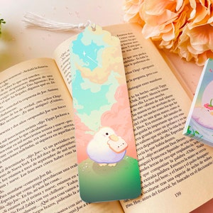 Puede incluir: Un marcapáginas colorido con un pájaro blanco con un pico rosa que sostiene un trozo de pan. El fondo es un cielo azul pastel, verde y rosa con nubes y estrellas. El marcapáginas tiene un borla.