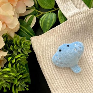 May include: A small, light blue plush stingray toy featuring embroidered black eyes and silver star-shaped accents on its upper surface. The soft, stuffed creature is positioned on a textured, off-white woven fabric surface.