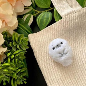 May include: A small, white, plush seal figurine with embroidered black eyes and a grey nose, attached to a beige, textured canvas fabric surface. The plush features a rounded body shape with simple grey stitched details representing its flippers.