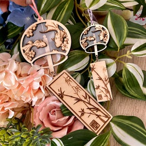 Puede incluir: Dos carillones de viento de madera con diseños intrincados. Uno presenta una campana circular con un patrón floral y siluetas de peces, mientras que el otro tiene una forma rectangular con un motivo de bambú. Ambos carillones están suspendidos de una cuerda.