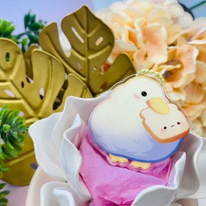 Puede incluir: Un llavero con un personaje de dibujos animados blanco y azul con una rebanada de pan amarilla. El personaje tiene un pequeño rubor rosa en las mejillas y sostiene la rebanada de pan en sus brazos. El llavero está sentado en una flor blanca y rosa.