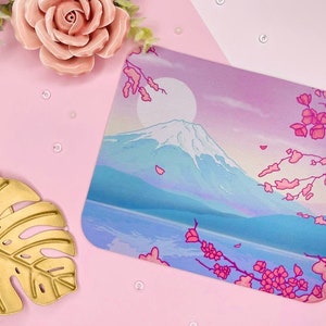 Può includere: Un tappetino per mouse quadrato con un paesaggio sereno raffigurante una montagna, un lago e fiori di ciliegio. Il design utilizza colori pastello blu, rosa e viola. Si vede una luna piena. Il tappetino è su una superficie rosa.