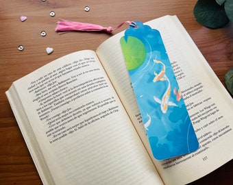 May include: A blue and green bookmark with a watercolor design of two koi fish swimming in a pond with a lily pad. The bookmark has a pink tassel.