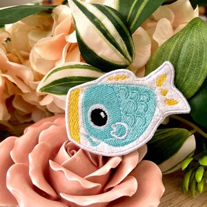 Puede incluir: Un parche bordado azul y blanco de un pez de dibujos animados con una aleta amarilla. El pez tiene un ojo grande y una boca pequeña.