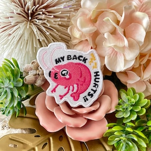 Puede incluir: Parche blanco bordado con una gamba rosa de dibujos animados y el texto "My Back Hurts"