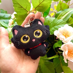 Puede incluir: Llavero de gato de peluche negro con ojos amarillos y un lazo rojo. El llavero se sostiene en una mano sobre un fondo verde de hojas.