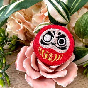 Puede incluir: Parche de hierro bordado rojo y blanco de un muñeco japonés Daruma con ojos negros y una inscripción amarilla. El parche está sobre una flor rosa.