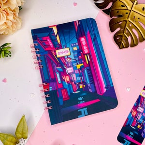 Puede incluir: Una libreta de espiral con una ilustración colorida de una calle de ciudad iluminada con luces de neón. La portada presenta caracteres japoneses y una combinación de colores rosa y azul brillantes.