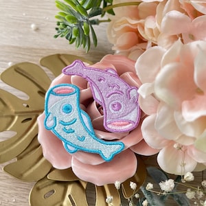 Puede incluir: Dos parches bordados para planchar con forma de pez. Un pez es azul claro con un ojo blanco y una boca rosa. El otro pez es morado con un ojo blanco y una boca rosa.
